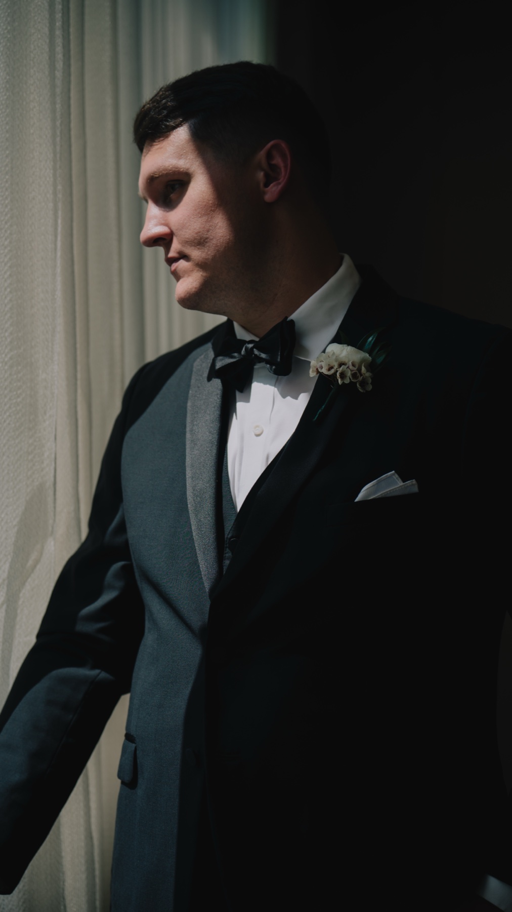 The groom in black tie waiting by a window in low light