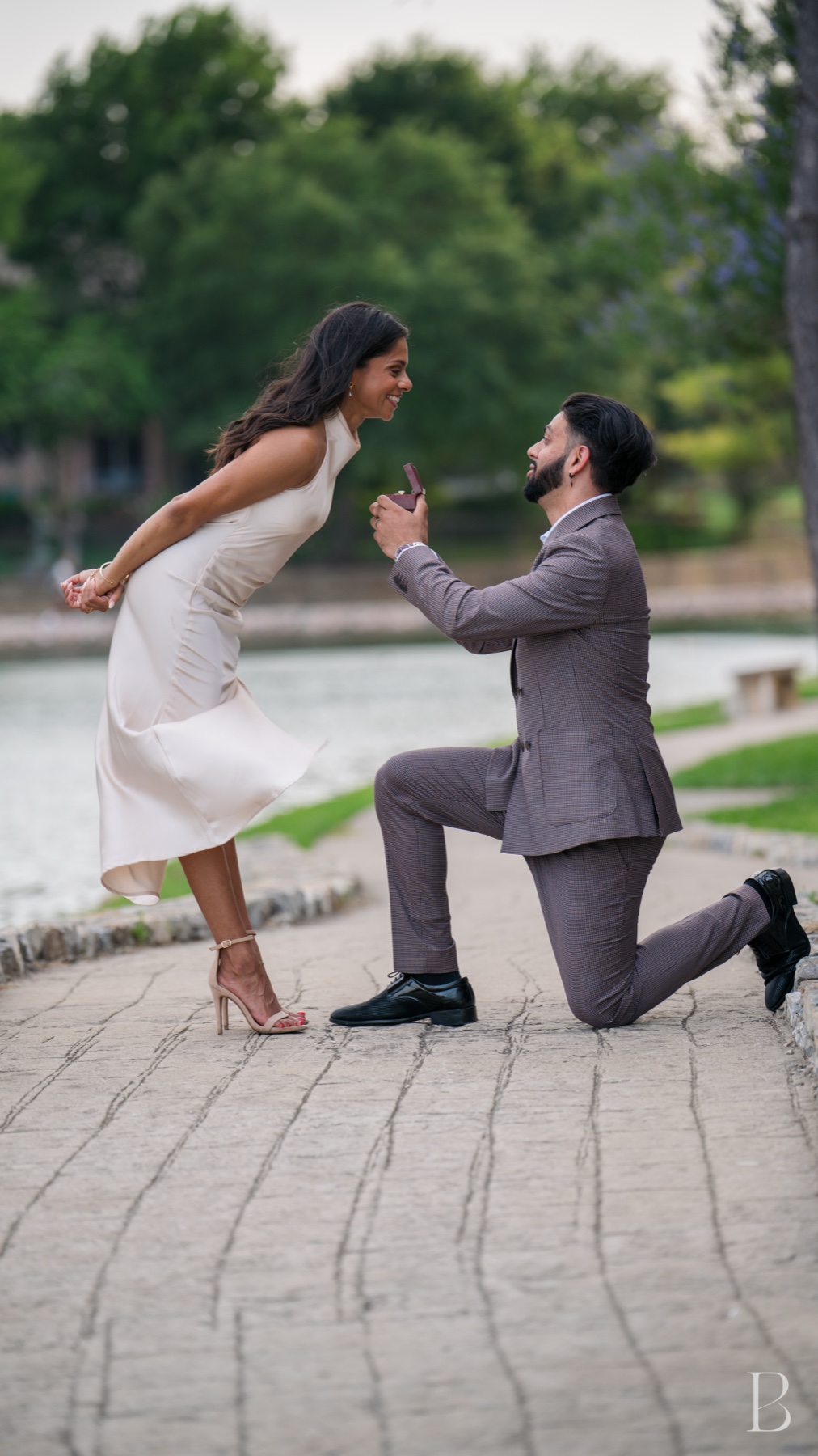 On one knee by the water, and she is laughing